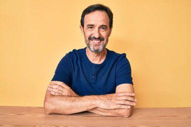 Orta yaşlı İspanyol bir adam, gündelik giysiler giyip masaya oturmuş kollarını kavuşturup kameraya bakıyordu. pozitif kişi. 