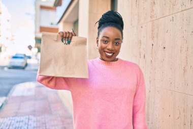 Genç Afro-Amerikalı kadın gülümseyerek elinde kağıt torbayla şehirde yiyecek taşıdığını söylüyor..