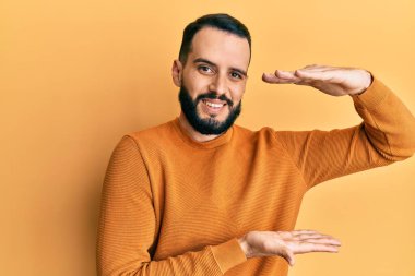 Sakallı, sıradan bir kazak giyen, elleri büyük ve büyük bir işaret gösteren, ölçü sembolü olan genç bir adam. Kameraya bakıp gülümsüyor. ölçüm kavramı. 