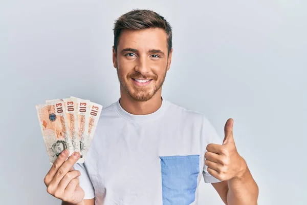 Handsome caucasian man holding 10 united kingdom pounds banknotes smiling happy and positive, thumb up doing excellent and approval sign