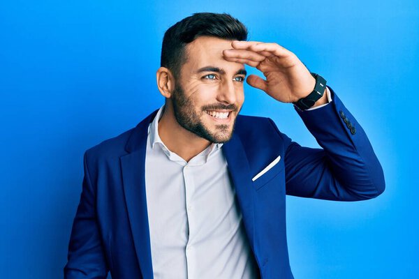 Young hispanic businessman wearing business jacket very happy and smiling looking far away with hand over head. searching concept. 