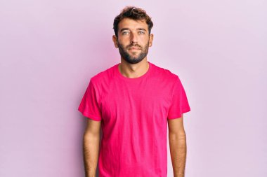 Yüzünde ciddi bir ifade olan pembe arka plan üzerine günlük pembe tişört giyen sakallı yakışıklı bir adam. Basit ve doğal bir şekilde kameraya bakıyorsun.. 