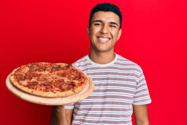 Genç Arap adam elinde İtalyan pizzası tutuyor. Pozitif ve mutlu görünüyor. Duruyor ve gülümsüyor. 