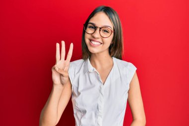 Genç esmer kadın, günlük kıyafetler ve gözlük takıyor. Üç numaralı parmaklarıyla gösteriyor ve işaret ediyor. Kendinden emin ve mutlu gülümsüyor.. 