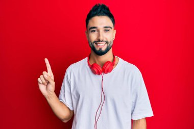 Sakallı genç adam müzik dinliyor. Yüzünde kocaman bir gülümseme olan kulaklıklar takıyor. Elinle kameraya bakıyor.. 