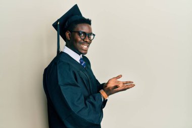 Mezuniyet şapkası ve tören cübbesi giyen yakışıklı siyahi adam avuçlarını açıp fotokopi alanını gösteriyor, gülümseyerek mutlu bir gülümseme sergiliyor. 