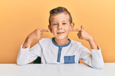 Gündelik giysiler giymiş sevimli beyaz çocuk masaya oturmuş neşeli bir şekilde gülümsüyor ve parmaklarıyla ve ağzıyla işaret ediyor. diş sağlığı kavramı. 