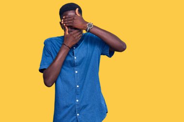 Genç afro-amerikan adam günlük kıyafetler giyiyor gözleri ve ağzı kapalı, şaşırmış ve şok olmuş. duygu gizleme 