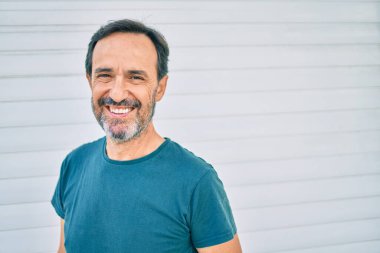 Orta yaşlı, sakallı, mutlu, açık havada duvara yaslanmış bir adam.