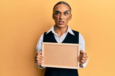 Makyaj yapan, uzun saçlı, boş mantar pano tutan, şüpheci ve gergin, sorun yüzünden kaşlarını çatan İspanyol bir adam. negatif kişi. 