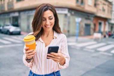 Genç İspanyol kadın mutlu bir şekilde gülümsüyor. Akıllı telefon kullanıyor ve şehirde kahve içiyor..