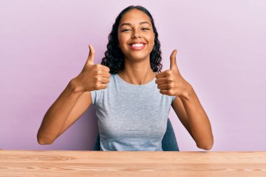 Genç Afro-Amerikan kızı gündelik giysiler içinde masada oturmuş, başarı işareti yapıyor, el hareketi yapıyor, gülümsüyor ve mutlu. Neşeli ifade ve kazanan jest. 