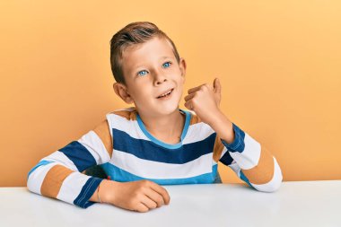 Sevimli beyaz çocuk, günlük giysiler içinde masaya oturmuş gülümsüyor, gülümsüyor ve başparmağıyla yan tarafa bakıyor.. 