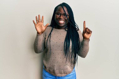 Saçları örgülü, günlük kıyafetler giyen ve gözlük takan genç siyahi bir kadın altı numarayı gösterip işaret ederken kendinden emin ve mutlu bir şekilde gülümsüyor.. 
