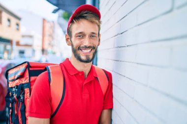 Beyaz teslimatçı, kırmızı üniforma giyiyor ve sırt çantası teslim ederken gülümsüyor.
