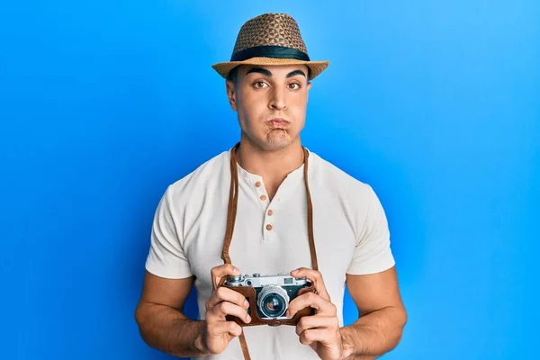 Hispanic young man holding vintage camera puffing cheeks with funny face. mouth inflated with air, catching air.
