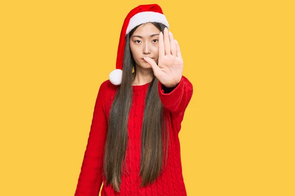 クリスマスの帽子をかぶっている若い中国の女性は手の手のひらで歌うのをやめます 顔に負の重大なジェスチャーで — ストック写真