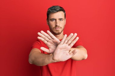 Yakışıklı, beyaz bir adam, günlük kırmızı tişört giyiyor. Reddedilmiş yüz ifadesi, kollar ve avuçlar üzerinde negatif işaretler, kızgın yüz ifadesi. 