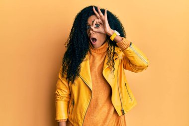 Orta yaşlı Afro-Amerikalı kadın yün kazak ve deri ceket giyiyor. Şaşırmış yüz ifadesiyle şaşırmış, parmaklarının arasından bakıyor. İnançsız ifade. 