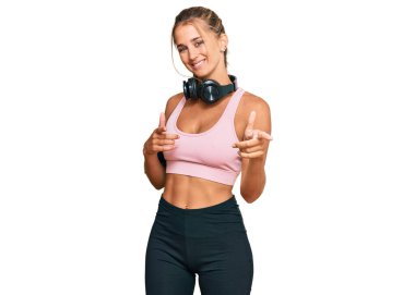 Spor kıyafetleri giyen ve kulaklık takan genç sarışın kadın kamerayı mutlu ve komik yüzüyle işaret ediyor. iyi enerji ve titreşimler. 