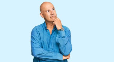 Orta yaşlı kel bir adam, elleri çenesinde gündelik giysiler giyip soru soruyor, düşünceli bir yüz ifadesi var. Gülümseyen ve düşünceli bir yüz. şüphe kavramı. 