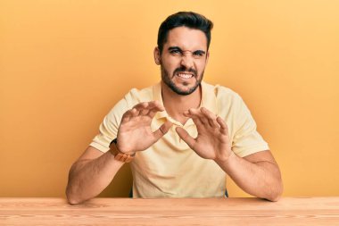 Latin kökenli genç bir adam, sıradan kıyafetler giymiş, tiksinmiş bir ifadeyle masada oturuyor, tiksinti tepkisi yüzünden tiksinti ve korku dolu bir ifade takınıyor. Eller havada. 