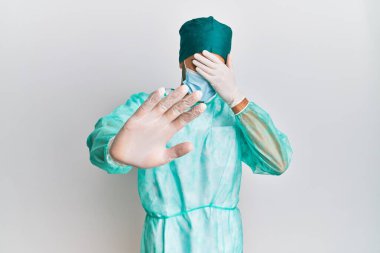 Cerrah üniforması giyen genç yakışıklı bir adam ve tıbbi maske takıyor ellerle gözleri örtüyor ve üzgün ve korku ifadeleriyle jestleri durduruyor. utangaç ve olumsuz kavram. 