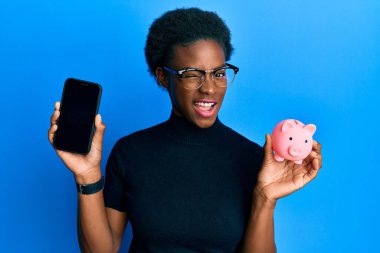 Genç Afrikalı Amerikalı kız domuz kumbarası tutuyor ve akıllı telefonuyla kameraya bakıyor. Seksi bir ifadeyle, neşeli ve mutlu bir yüzle.. 