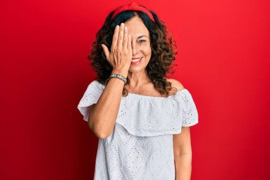 Orta yaşlı İspanyol kadın gündelik kıyafetler giyiyor. Bir gözünü eliyle kapatıyor. Yüzünde kendine güvenen bir gülümseme ve sürpriz bir duygu var.. 