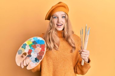 Güzel beyaz kız elinde boya fırçası ve ressam paleti ile çılgınlığı kutluyor ve başarı için hayrete düşüyor.. 