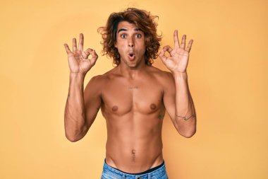 Genç İspanyol adam tişörtsüz duruyor. Şaşırmış ve şok olmuş görünüyor. Tamam sembolünü parmaklarıyla yapıyor. çılgın ifade 