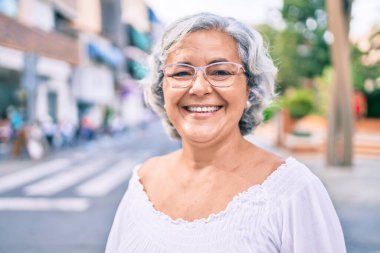 Beyaz saçlı orta yaşlı bir kadın mutlu bir şekilde dışarıda gülümsüyor.