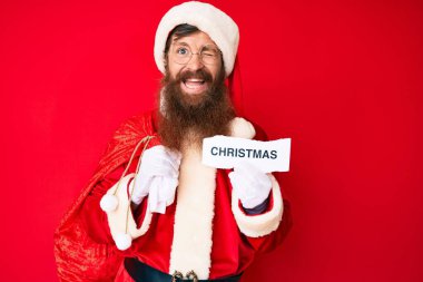 Uzun sakallı, yakışıklı, Noel Baba kostümü giyen, elinde noel mesajı tutan, seksi bir ifadeyle kameraya bakan, neşeli ve mutlu yüzlü genç bir adam.. 