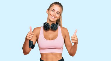 Spor kıyafetleri giyen, kulaklık takan, el hareketleriyle olumlu jest yapan, gülümseyen ve mutlu genç sarışın bir kadın. Neşeli ifade ve kazanan jest. 