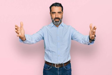 Orta yaşlı, sakallı, iş gömleği giyen, kameraya bakıp sarılmak için kollarını açmış gülümseyen bir adam. Mutluluğu kucaklayan neşeli bir ifade.. 