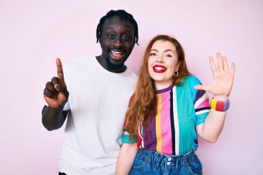 Irklar arası çift gündelik kıyafetler giyer ve altı numaralı parmaklarıyla gülümseyerek mutlu ve kendinden emin bir şekilde işaret eder.. 