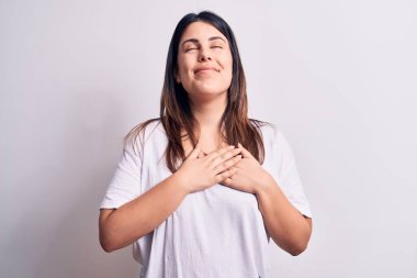 Genç, güzel, esmer, günlük tişört giyen bir kadın izole edilmiş beyaz arka planda duruyor. Elleri göğsünde, gözleri kapalı, yüzünde minnet dolu bir jestle gülümsüyor. Sağlık kavramı.