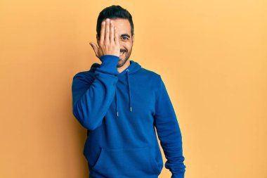 Latin asıllı genç adam gündelik giysiler giyiyor. Bir gözünü eliyle kapatıyor. Yüzünde kendine güvenen bir gülümseme ve sürpriz bir duygu var.. 