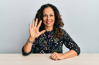 Masada oturan orta yaşlı, güzel bir kadın beş numaralı parmaklarını gösterip gülümserken kendinden emin ve mutlu görünüyor.. 