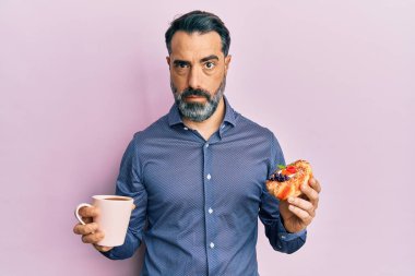 Orta yaşlı, sakallı, kır saçlı, kahve içen, hamur işi yiyen, şüpheci ve gergin bir adam sorun yüzünden kaşlarını çatıyor. negatif kişi. 