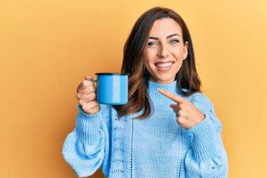 Genç esmer kadın elinde kahve fincanı tutarken mutlu bir şekilde gülümsüyor. 