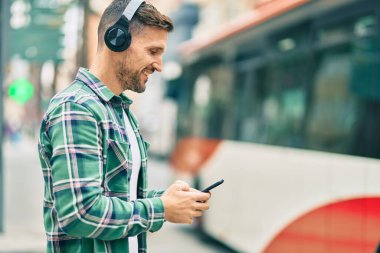 Genç beyaz adam şehirde akıllı telefon ve kulaklık kullanarak mutlu bir şekilde gülümsüyor..