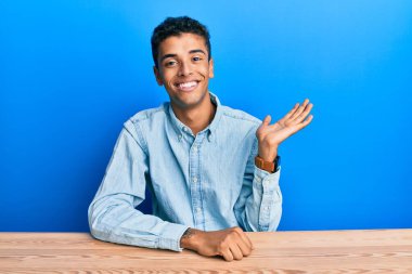 Genç, yakışıklı, Afro-Amerikalı, günlük giysiler içinde masaya oturmuş neşeli bir şekilde gülümsüyor ve avuç içi kameraya bakıyor.. 