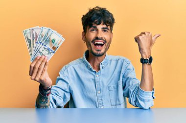 Masada dolarları tutan Latin asıllı genç bir adam başparmağıyla yan tarafı işaret ediyor ve ağzı açık gülümsüyor. 