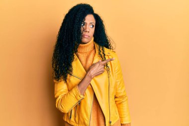 Middle age african american woman wearing wool winter sweater and leather jacket pointing with hand finger to the side showing advertisement, serious and calm face 