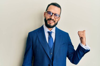 Sakallı, takım elbiseli, kravatlı, mutlu yüzlü, gülümseyen ve baş parmağıyla yan tarafı gösteren genç bir adam.. 