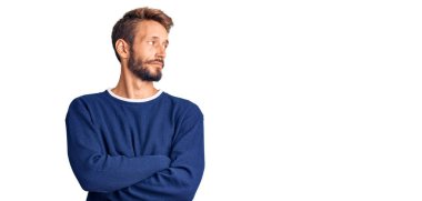 Sarışın, sakallı, sıradan bir süveter giyen kolları çapraz ve kendinden emin bir şekilde yana bakan yakışıklı bir adam. 