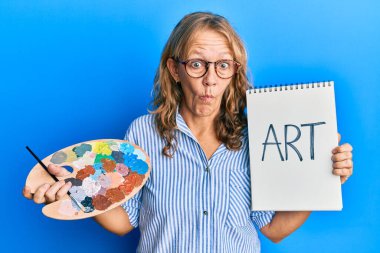 Orta yaşlı sarışın kadın boyacı paleti ve not defteri tutuyor. Ağzı ve şaşı gözleriyle balık suratı yapıyor. Deli ve komik.. 