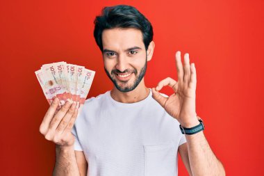 Young hispanic man holding 10 colombian pesos banknotes doing ok sign with fingers, smiling friendly gesturing excellent symbol 