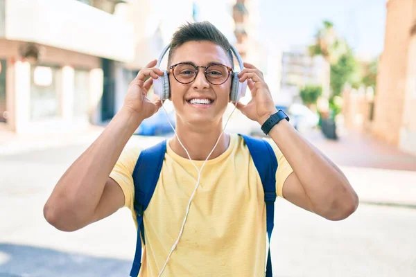 Young hispanic student smiling happy using headphones walking at street of city